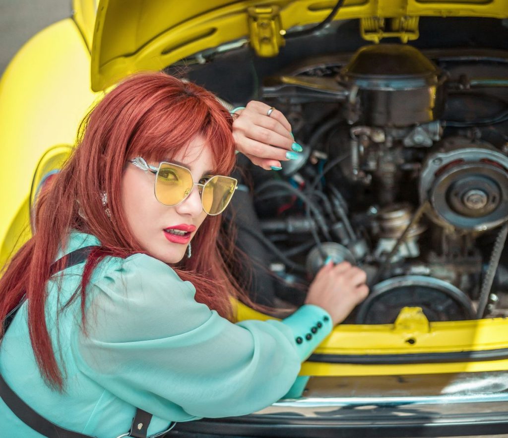 Young woman dressed in a blouse with sunglasses, leaning over an open hood of a yellow car.