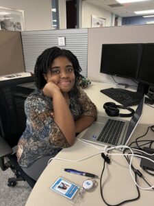 Damikka Willis, Marketing Intern at MOTOR, sitting at her office desk with a laptop, smiling and resting her chin on her hand, with MOTOR badge and pen visible.