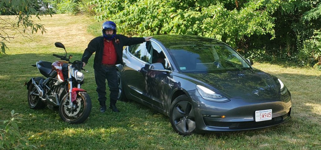 Craig Van Batenburg standing outside between a motorcycle and Tesla.
