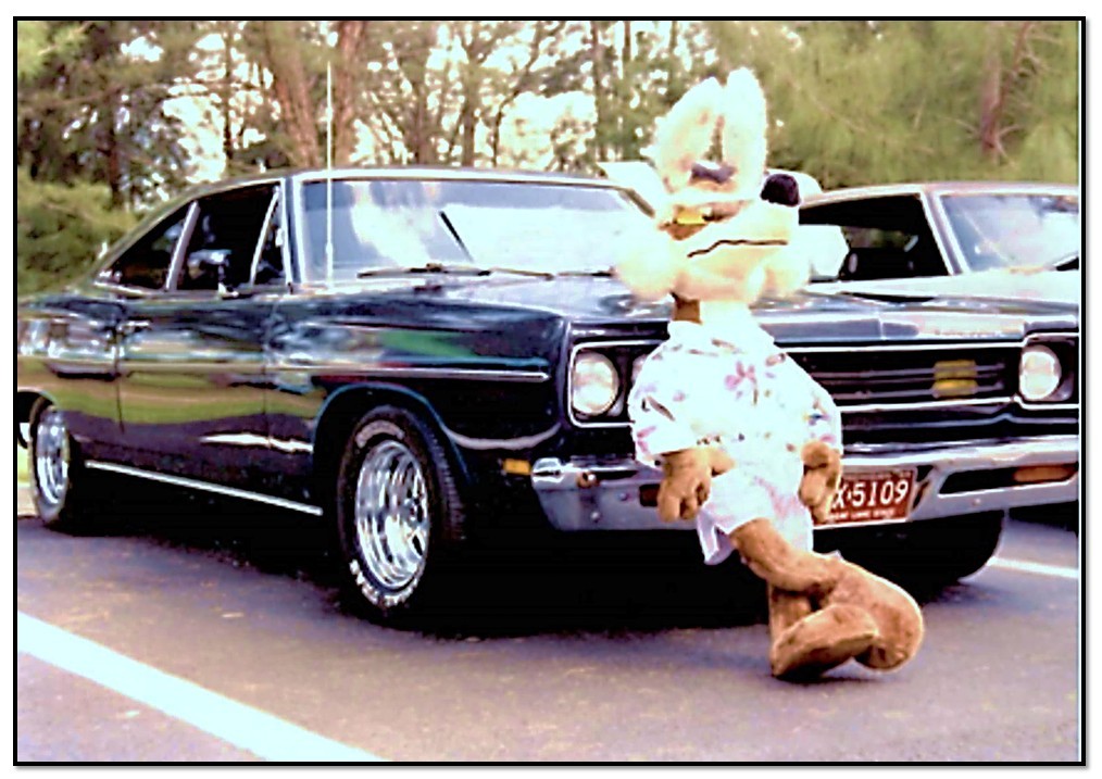 A large stuffed mascot character in a tropical shirt and white shorts leaning against a classic dark blue muscle car parked outdoors. The car has chrome wheels and a visible license plate reading "XK5109". Another similar car is parked beside it.