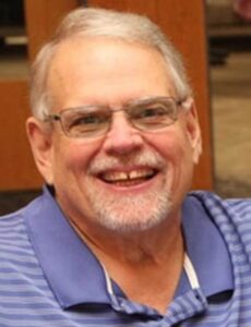 Portrait of an older man smiling and wearing glasses and a blue striped polo shirt, seated indoors with a blurred background.