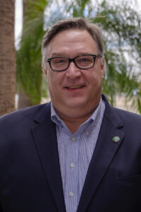 Portrait of a middle-aged man wearing black glasses, a navy blazer with a green lapel pin, and a blue-striped button-down shirt, standing outdoors with palm trees in the background.