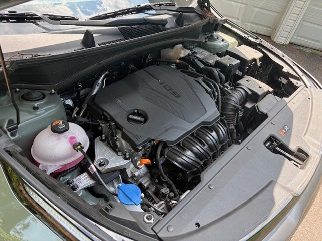 View of a modern vehicle engine bay with a GDI (Gasoline Direct Injection) engine cover, showing various engine components including the coolant reservoir, oil cap, battery, and windshield washer fluid reservoir. The hood is open in a residential driveway, with part of a garage visible in the background.