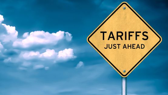 Yellow diamond-shaped road sign with the word “TARIFFS” against a blue sky with clouds, symbolizing trade policies and economic impact.