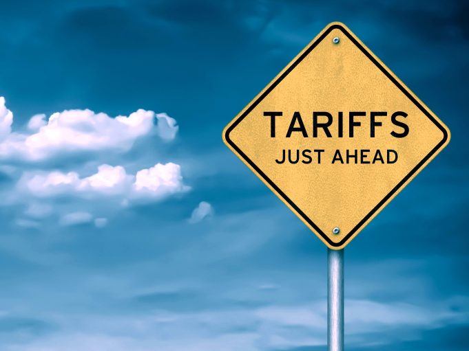 Yellow diamond-shaped road sign with the word “TARIFFS” against a blue sky with clouds, symbolizing trade policies and economic impact.