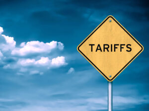 Yellow diamond-shaped road sign with the word “TARIFFS” against a blue sky with clouds, symbolizing trade policies and economic impact.